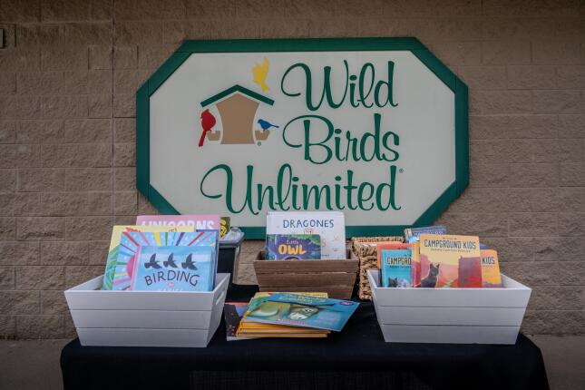 Colorful children&rsquo;s books arranged in baskets and crates on a table beneath the Wild Birds Unlimited sign, ready to be given away for Dolly Parton Day.